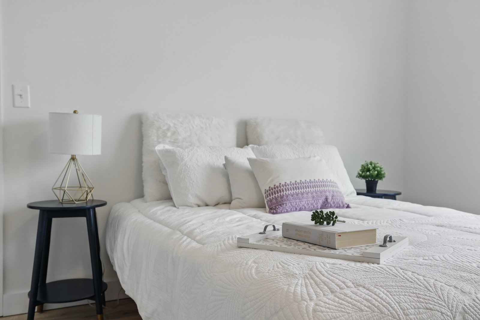 Staged bedroom in soft natural light with white linens and a book tray